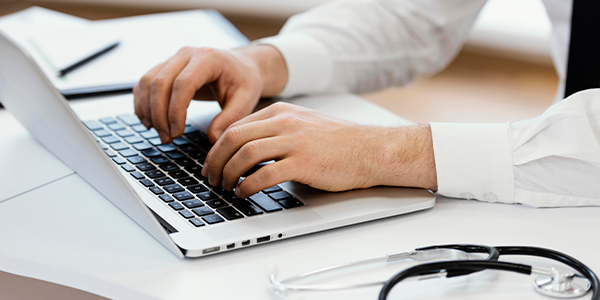 Mãos de médico teclando em notebook aberto em agenda médica online.